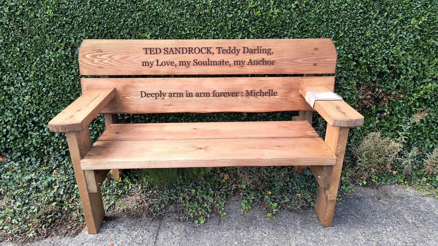Memorial bench with carved lettering, photographed against a hedge